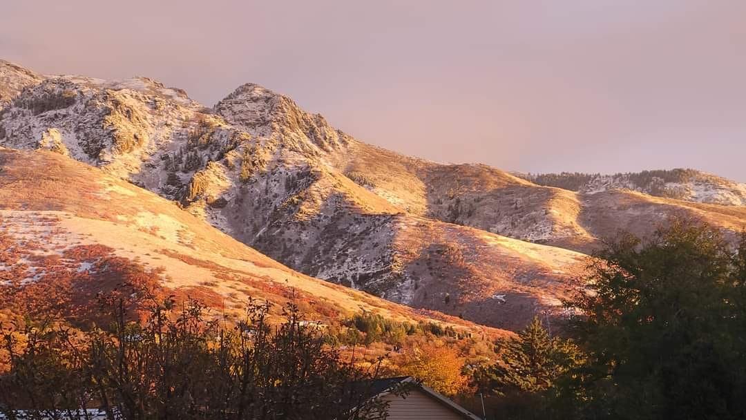 Golden sunset light illuminates snow-dusted mountain peaks and reddish slopes above a residential valley.