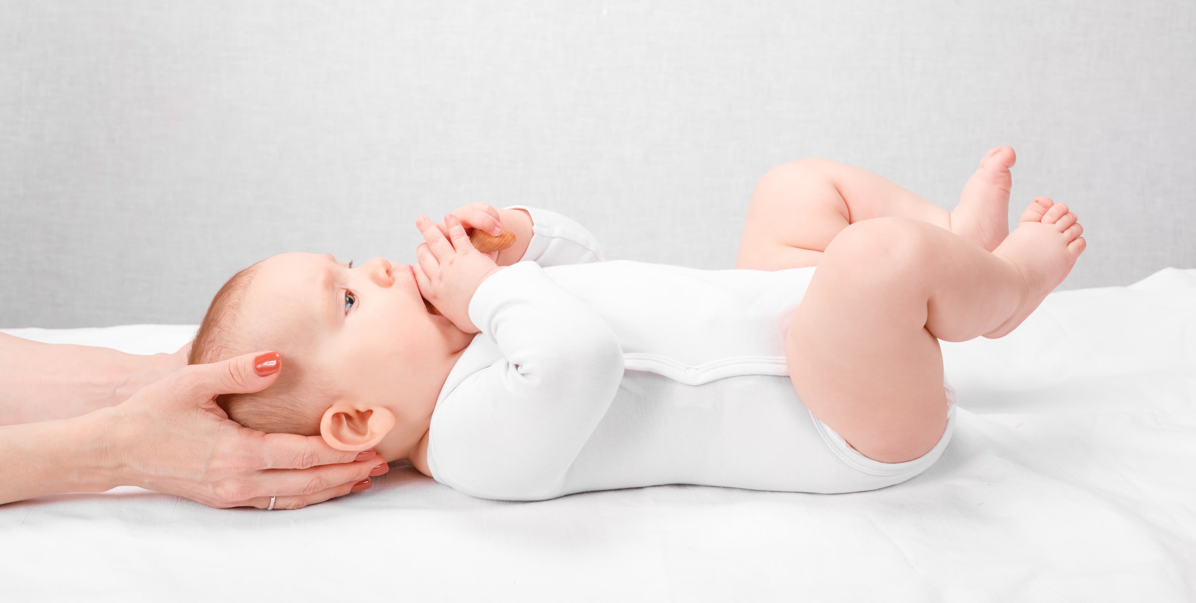 Infant in a white bodysuit lies on its back while hands gently cradle its head.