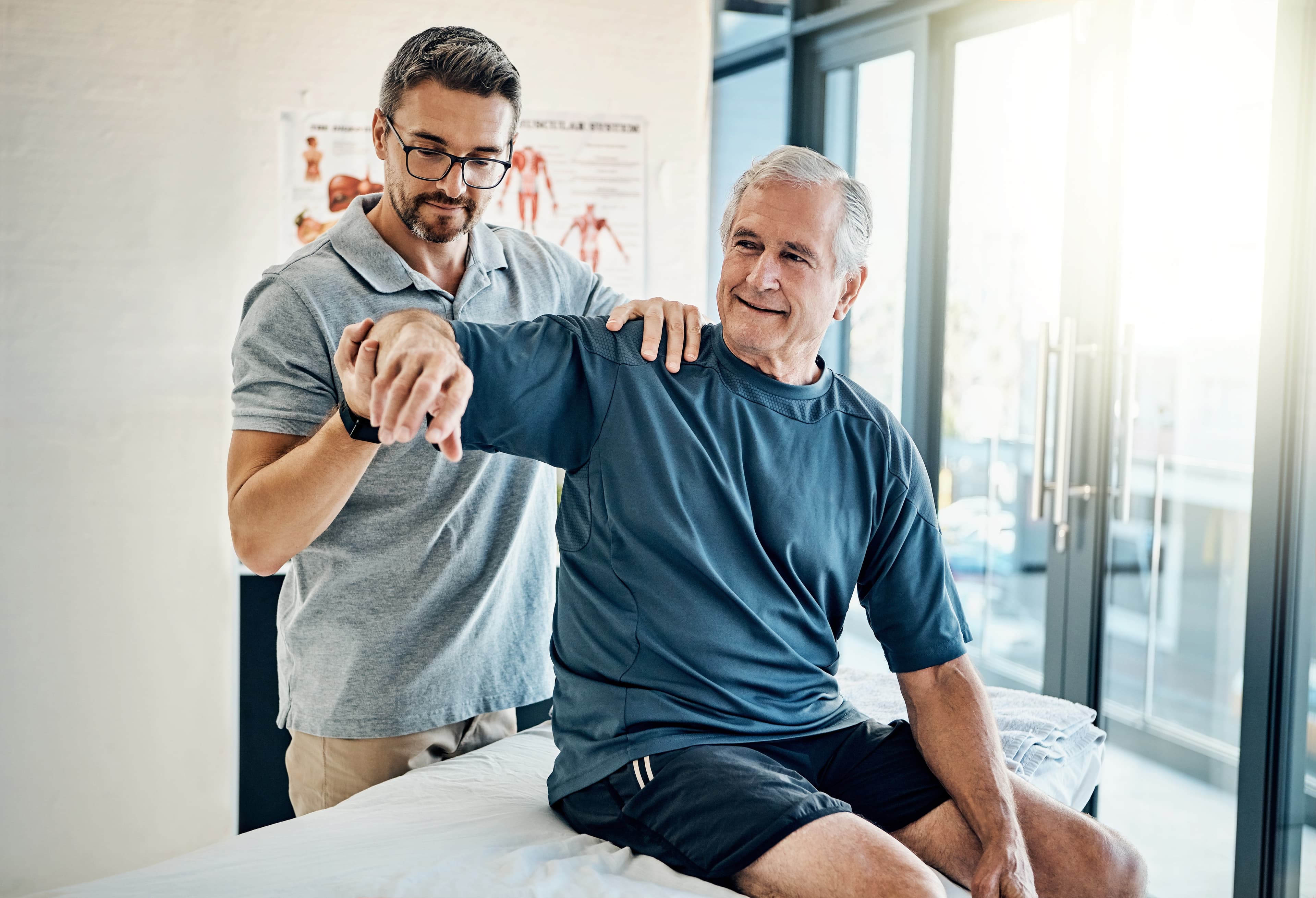Male physical therapist assisting an elderly patient with shoulder mobility exercises in a bright clinic.